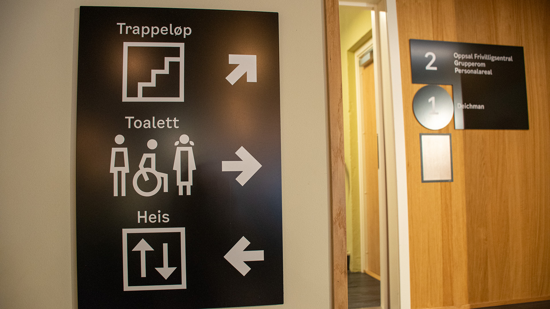 Tre skyltar på eb vit vägg. Med både bilder, text och pilar visar de vägen till toalett, trappor och hiss. 