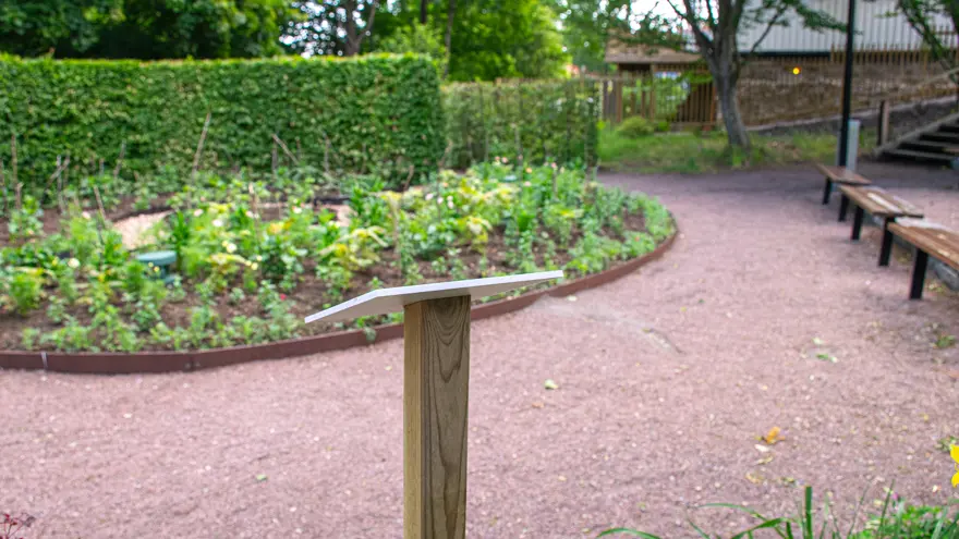I förgrunden står en mindre skylt på en trästolpe. Den står vid en grusgång och längre bort finns en rabatt och bänkar.