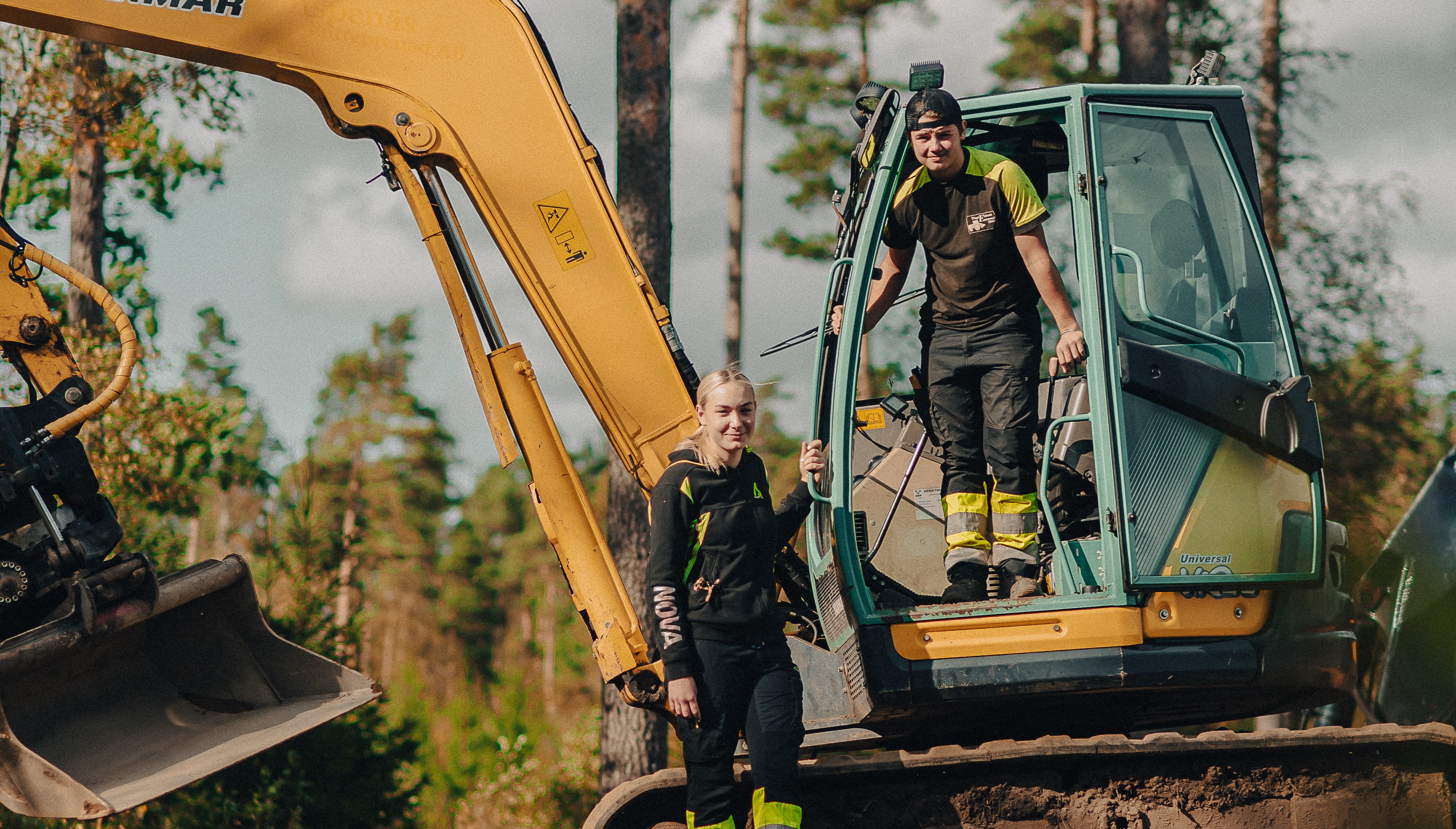 Två elever står vid grävmaskin i skogen