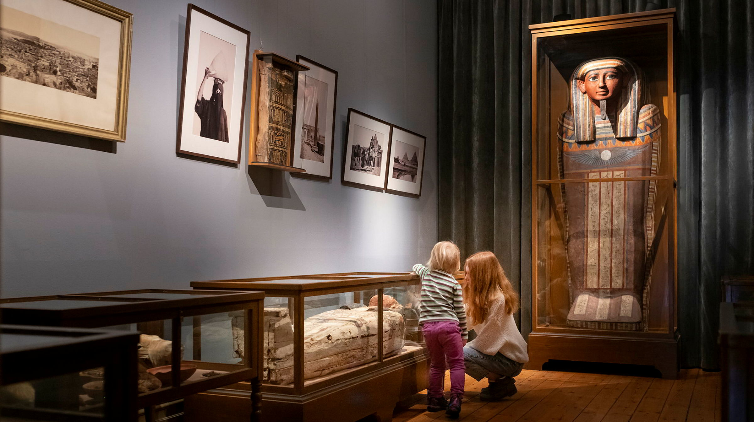 Två barn syns titta på en sarkofag till en mumie i en glasmonter, bakom dem syns en annan egyptisk sarkofag. Äldre museimiljö.