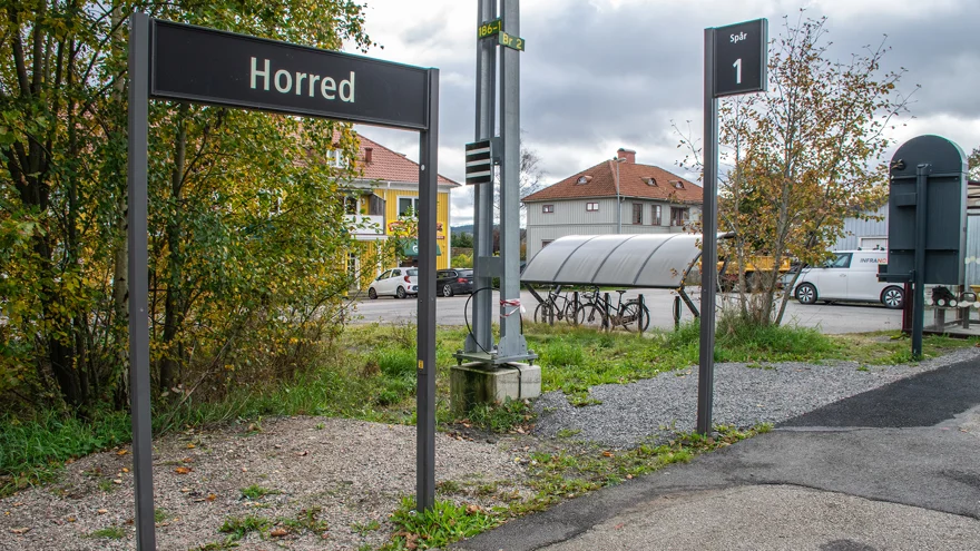 En skylt med texten Horred står på en tågperrong. bakom skylten finns ett par trähus och ett cykelställ.