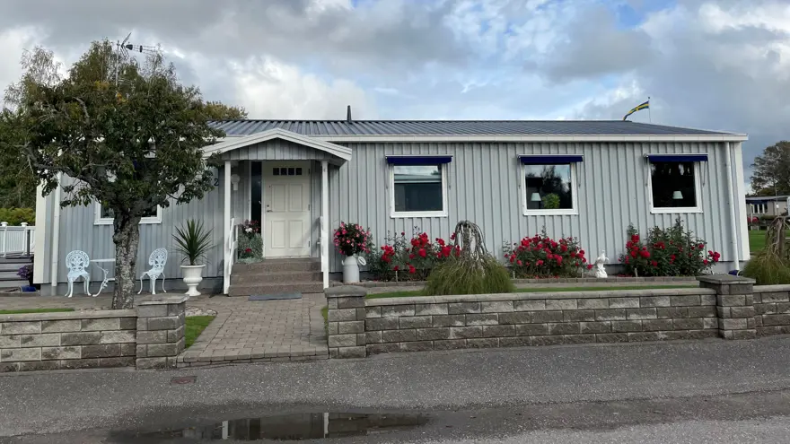 Ett ljusgrått enplanshus med en stenlagd gårdsplan framför. Till vänster är ett litet träd framför huset och till höger många blommor i rött och rosa.