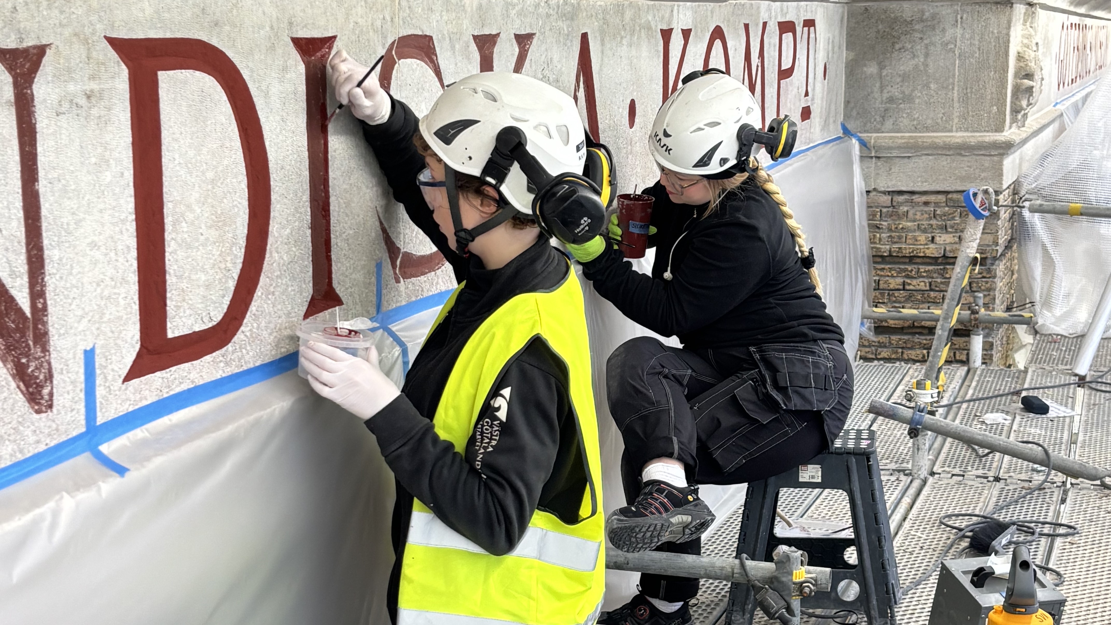 En stenfasad med röda bokstäver infasade. Två kvinnor arbetar med att fylla i bokstäverna med röd färg. De är klädda i bygghjälmar samt arbetskläder, eom en gul reflexväst närmast kameran. 