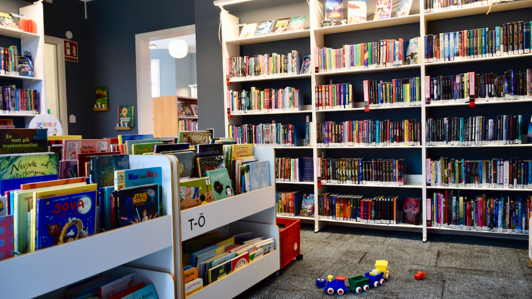 Barnavdelningen på biblioteket. På golvet finns en heltäckningsmatta, väggarna är målade i en dov gråblå färg. Bokhyllor utmed väggarna och boklådor i mitten av rummet och på golvet står ett färgglatt leksakståg i plast. 