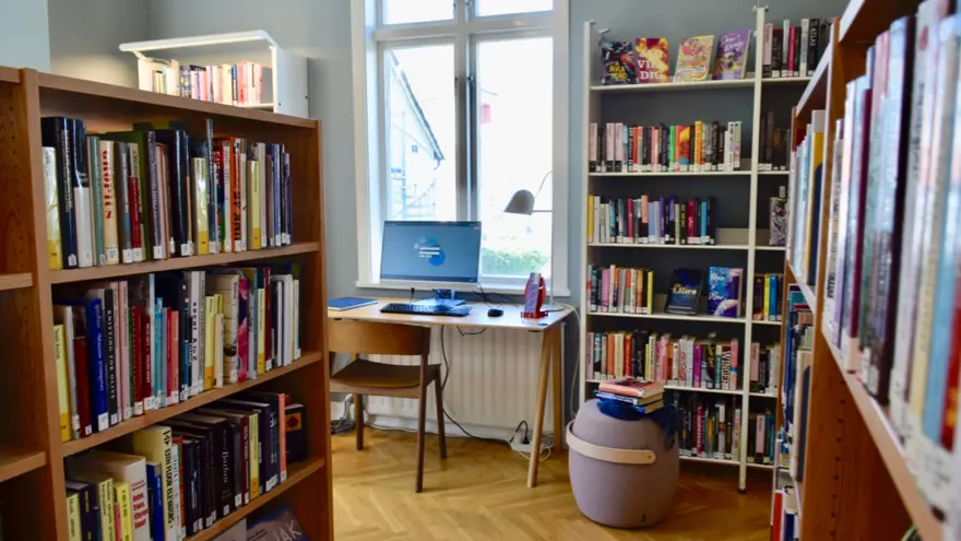 Fyllda bokhyllor inne på vuxenavdelningen. Vid ett fönster står ett litet skrivbord med en datorskärm och en stol.