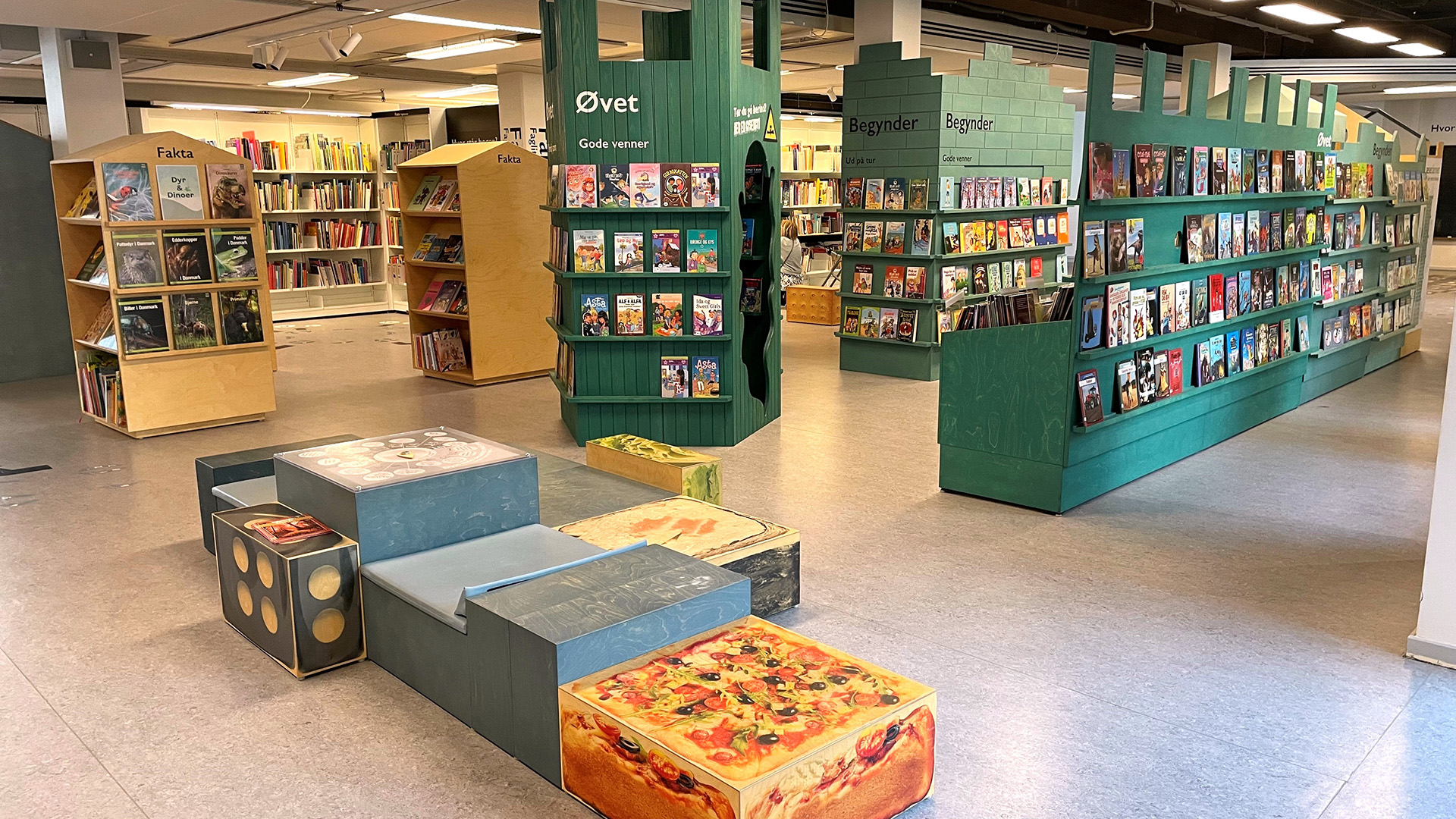 Barnavdelningen LÄS, interiör. Bokhyllor i grönt och ljust trä, böckerna är i många fall placerade med framsidan utåt. En sittmöbel finns mitt i rummet, stora sittytor i olika nivåer. Golvet har en beige plastmatta.