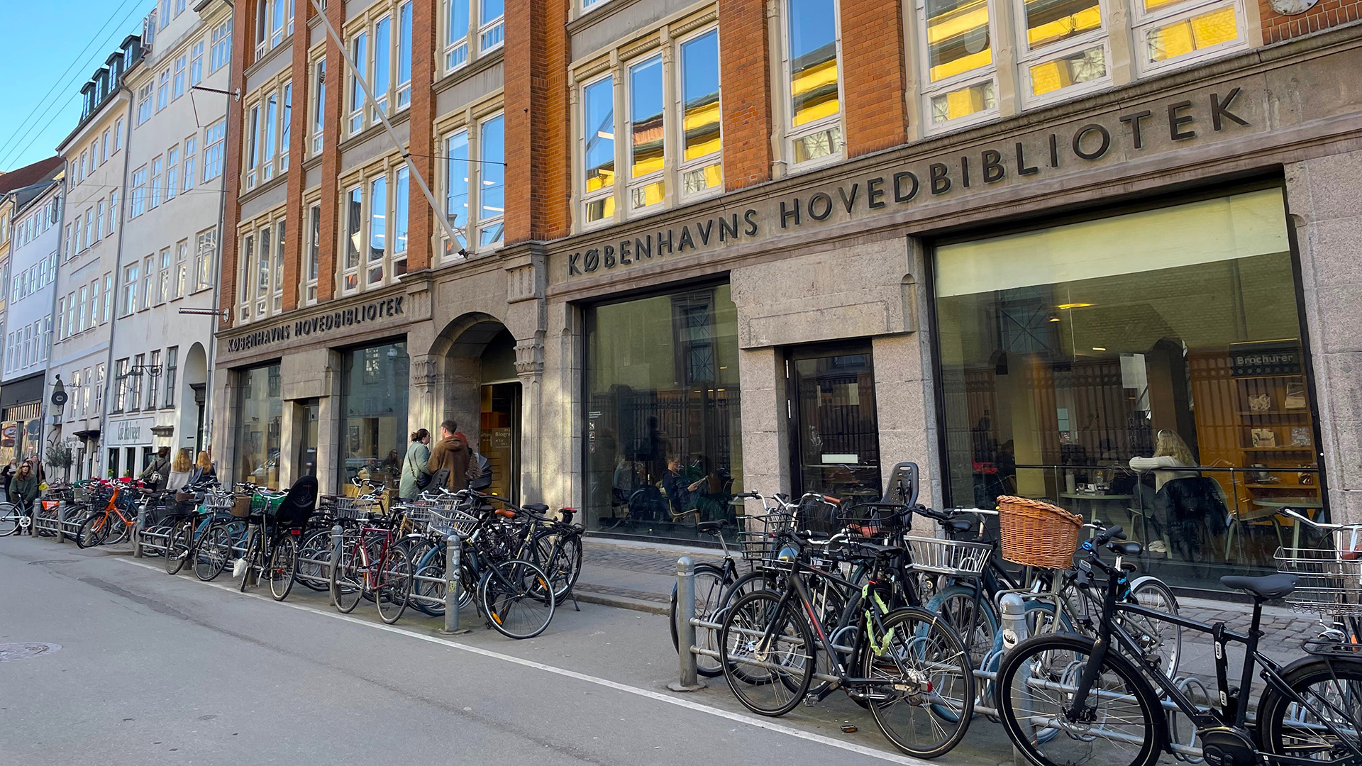 En gata i Köpenhamn. En byggnad med stora skyltfönster mot gatan, utanför står många cyklar parkerade. Ovanför ingången står det med stora bokstäver KÖPENHAMNS HUVUDBIBLIOTEK på danska. 