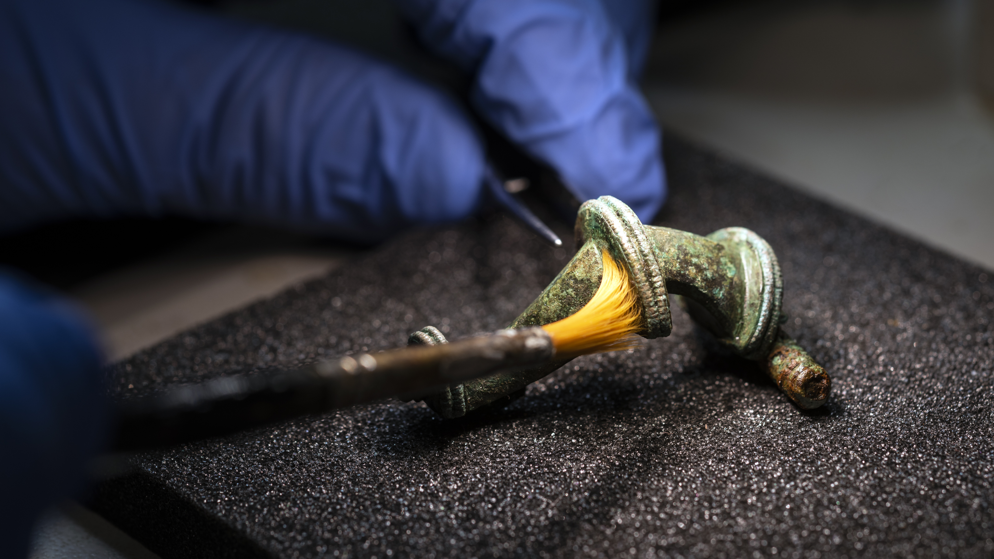 En hand med blå latexhandske penslar försiktigt på ett litet ömtåligt metallföremål.